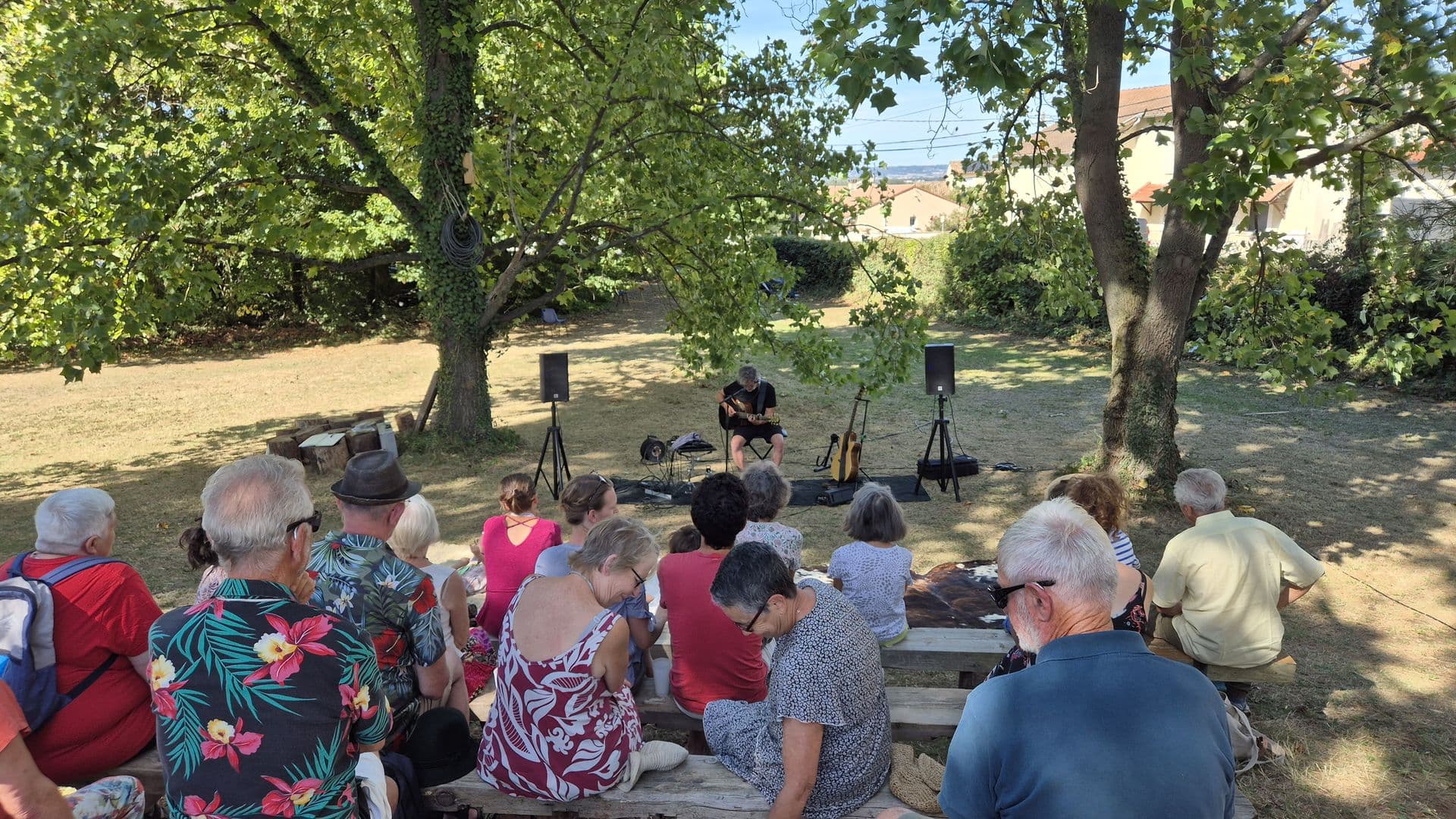 Vue depuis l’arrière du public : le musicien seul entre ses enceintes dans le parc de la Maison Pérouze, et les toits du village au loin