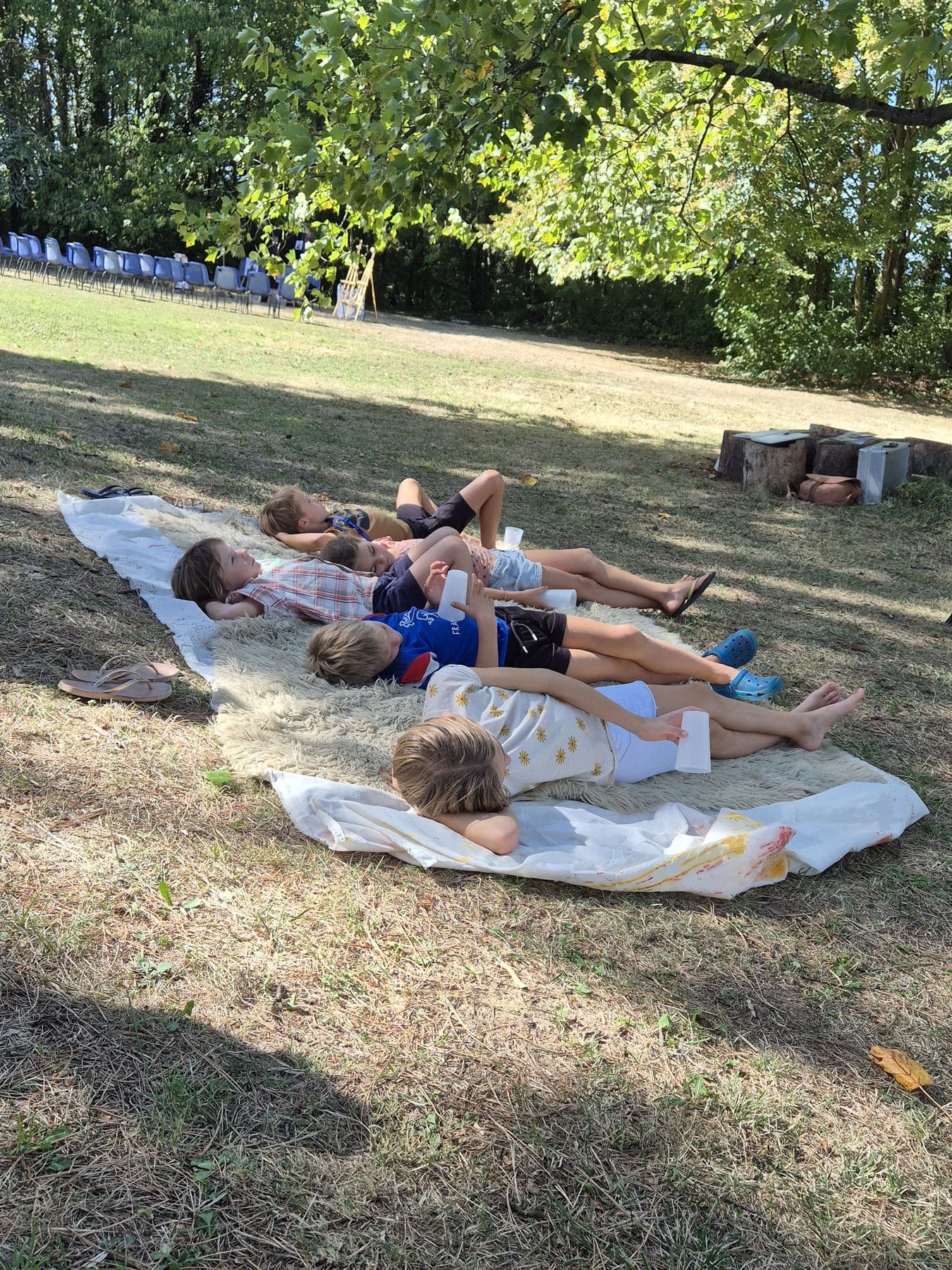 Des enfants allongés sur des tapis, à l’ombre, un peu à l’écart des bancs