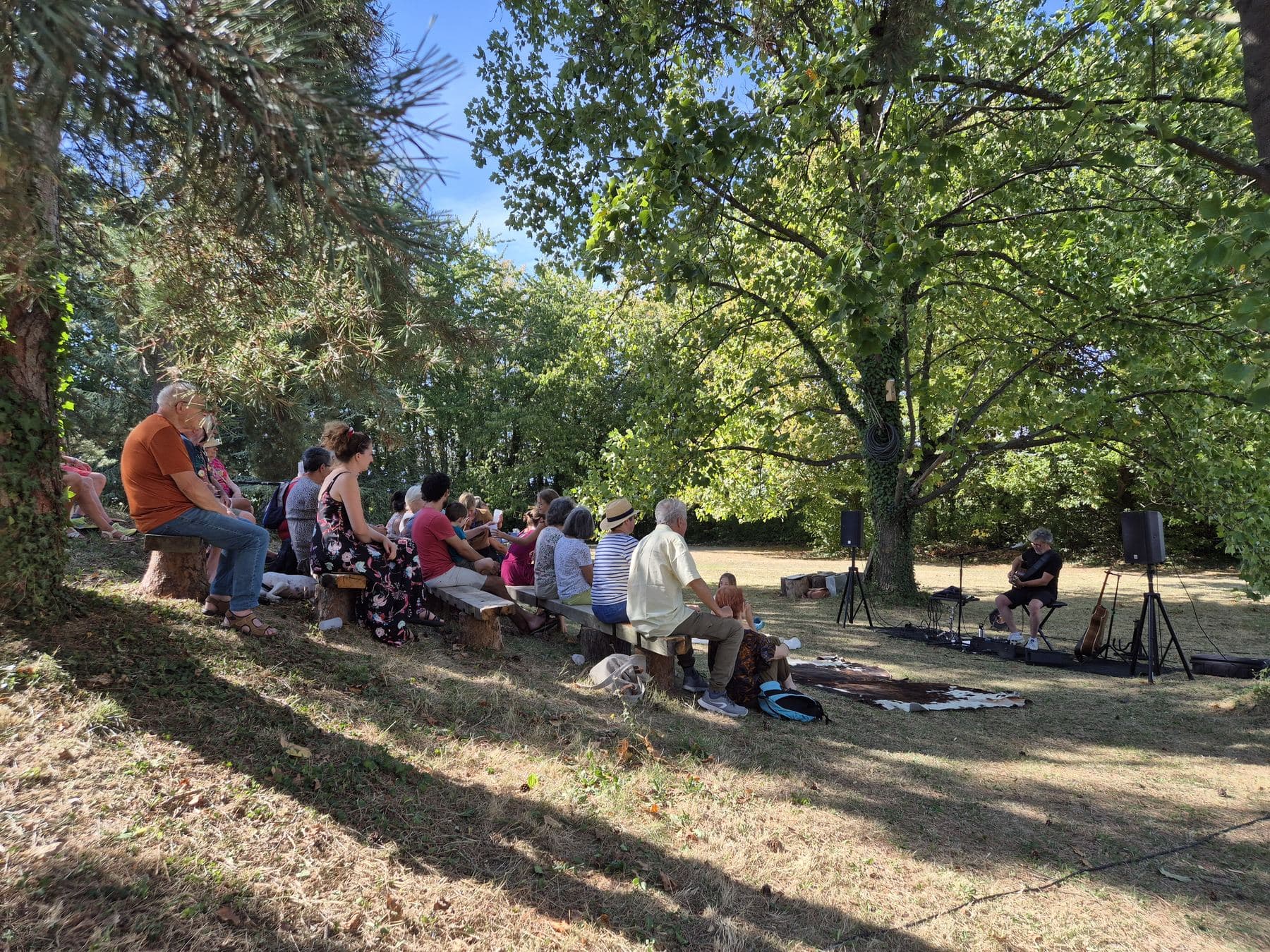 Le public assis sur les bancs de bois, à flanc de talus sous les grands arbres du parc, face au musicien